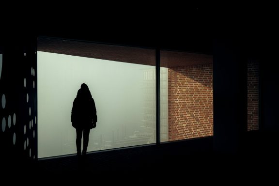 Eine Person steht silhouettiert hinter einer großen Fensterfront in nebliger Umgebung.
